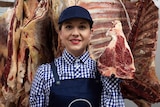 A smiling woman holding a cut of meat, carcass and meat hanging behind her.