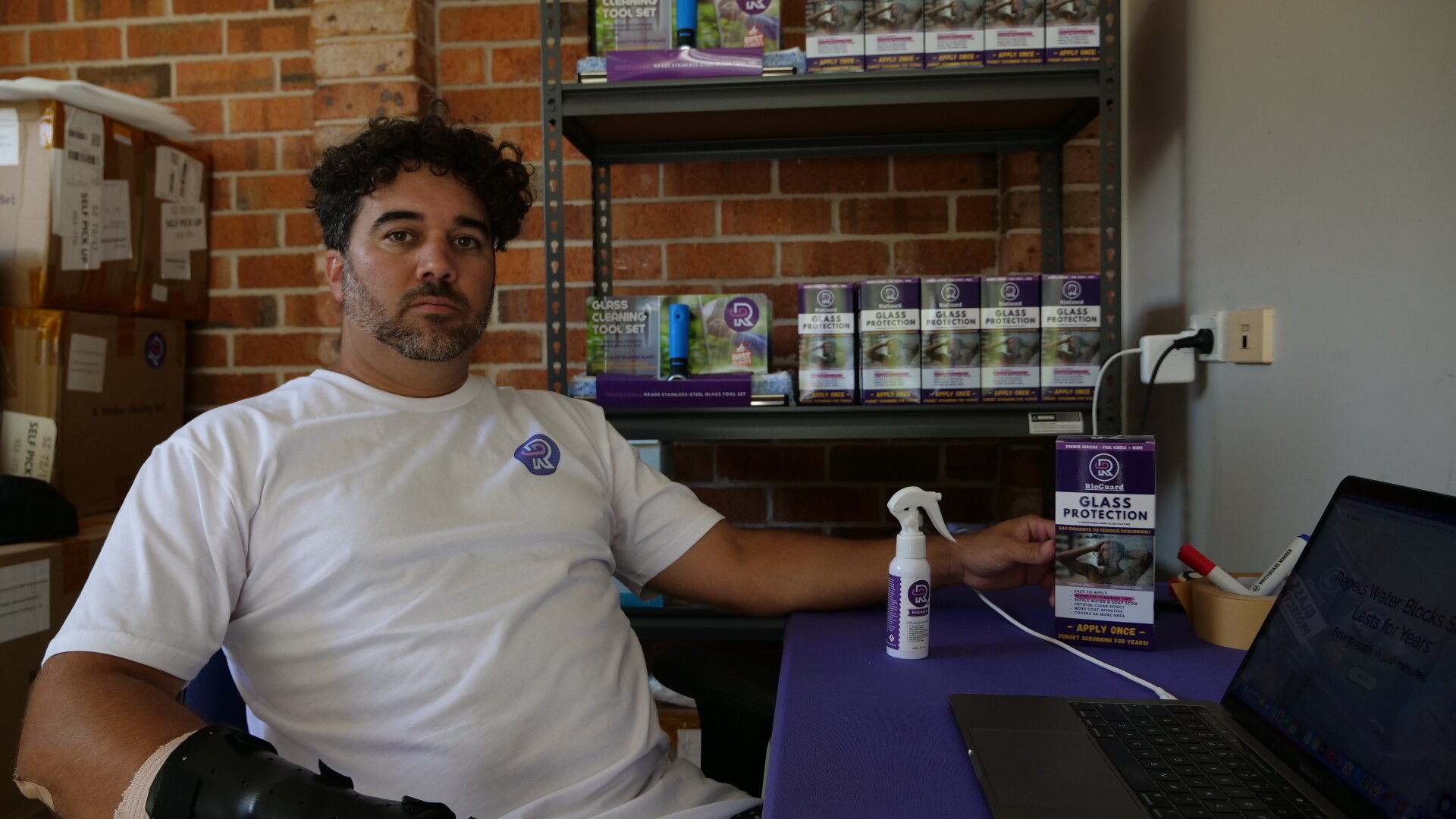 Um jovem de cabelos escuros e encaracolados está sentado em uma mesa no que parece ser uma garagem.