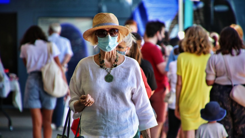 A woman in a sun hat, linen top and surgical face mask walks through a crowd on a sunny day.