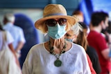 A woman in a sun hat, linen top and surgical face mask walks through a crowd on a sunny day.