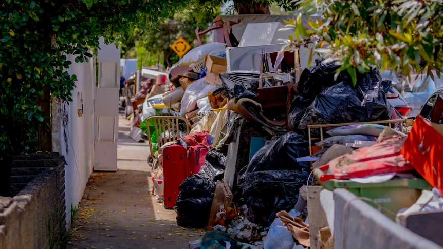 A suburban street. On one side is the fence of a house, on the other are piles of ruined houshold items