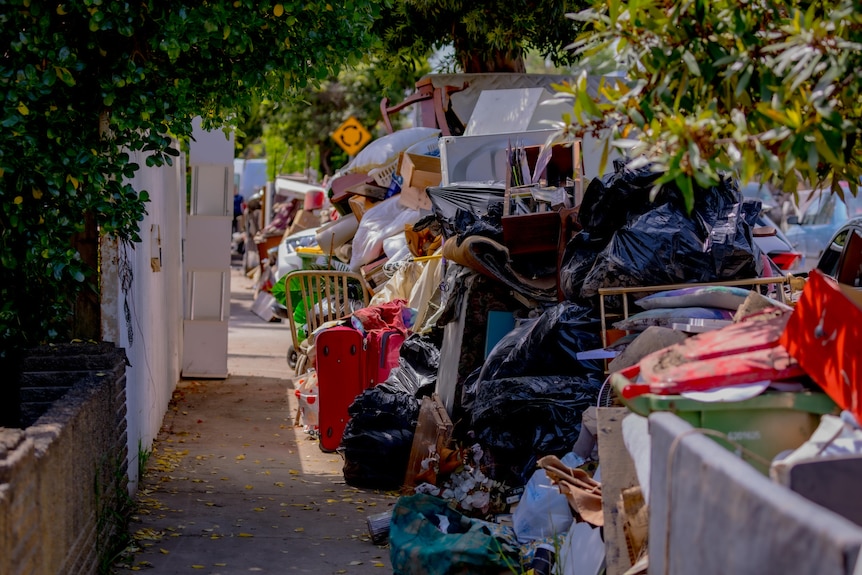 Une rue de banlieue.  D'un côté se trouve la clôture d'une maison, de l'autre des tas d'objets ménagers en ruine