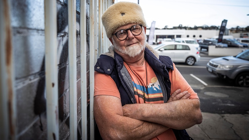 A man with a white beard wearing an orange t-shirt and a jacket leans on a car park fence.