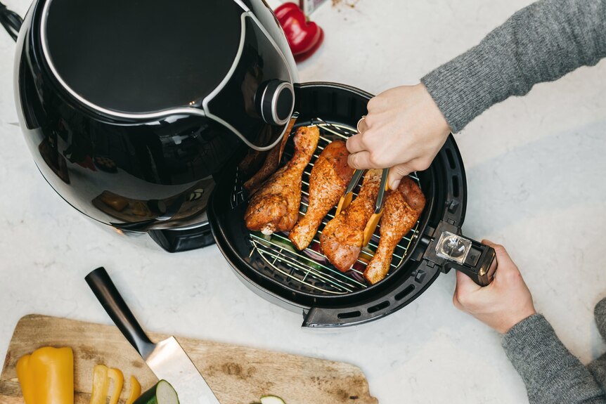 A person in a grey jumper uses tongs to turn chicken drumsticks in a black air fryer.