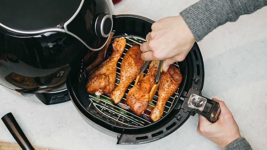 A person in a grey jumper uses tongs to turn chicken drumsticks in black air fryer, in a story about the best air fryer recipes