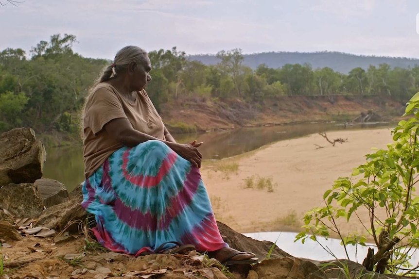 Older Indigenous woman sits on a rock and looks out into the Northern Territory landscape.