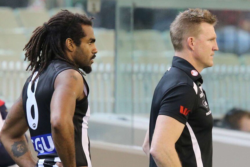 Collingwood coach Nathan Buckley stands in front of Héritier Lumumba on the sideline of an AFL game.