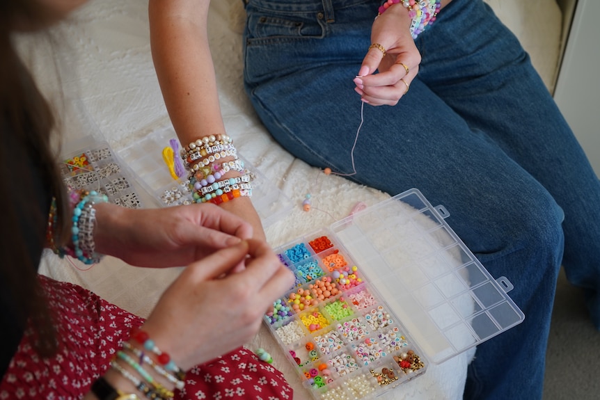 Two women making beaded friendship bracelets on a bed