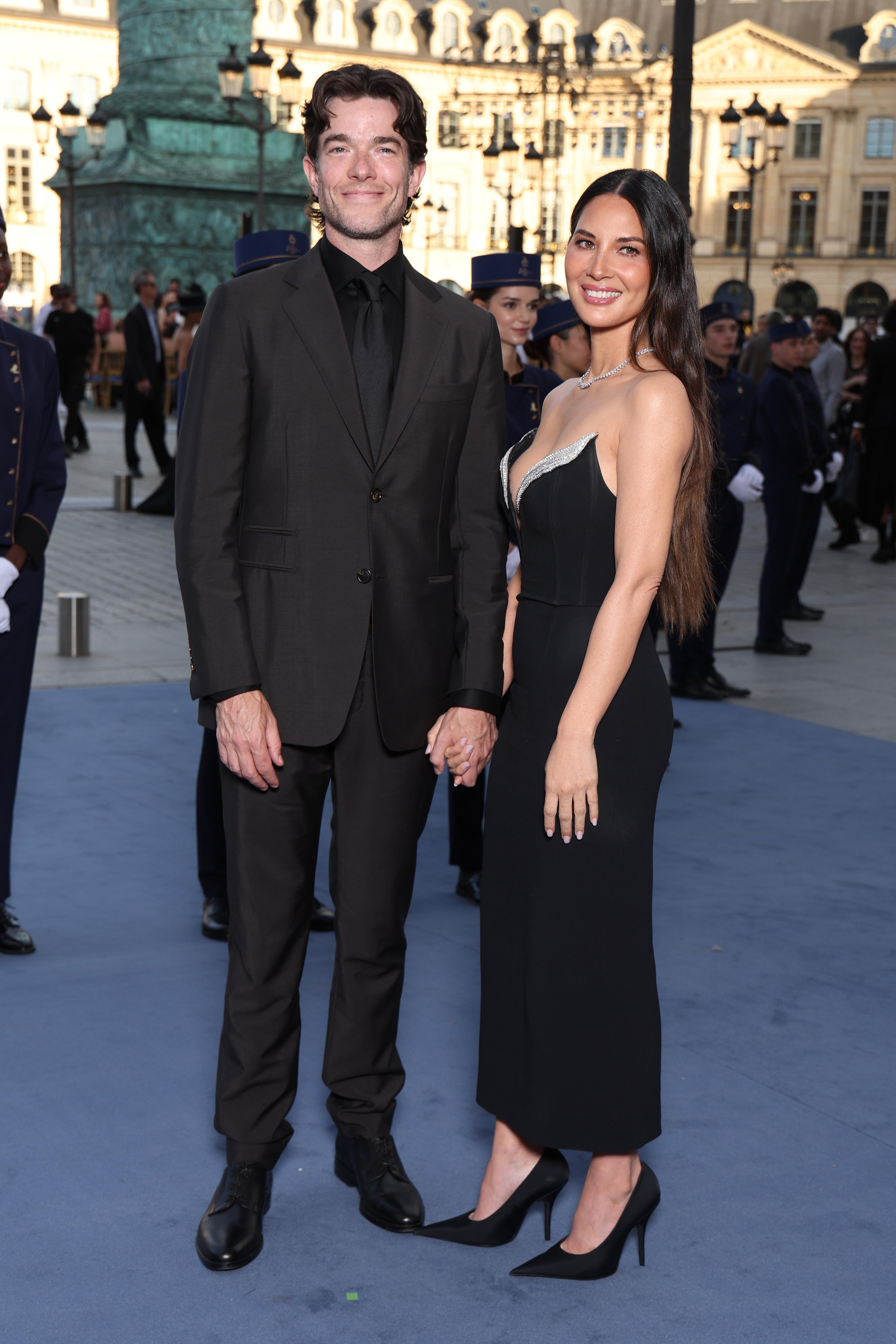 John Mulaney, in a black suit, and Olivia Munn, in a black dress and heels, smile for a photo.
