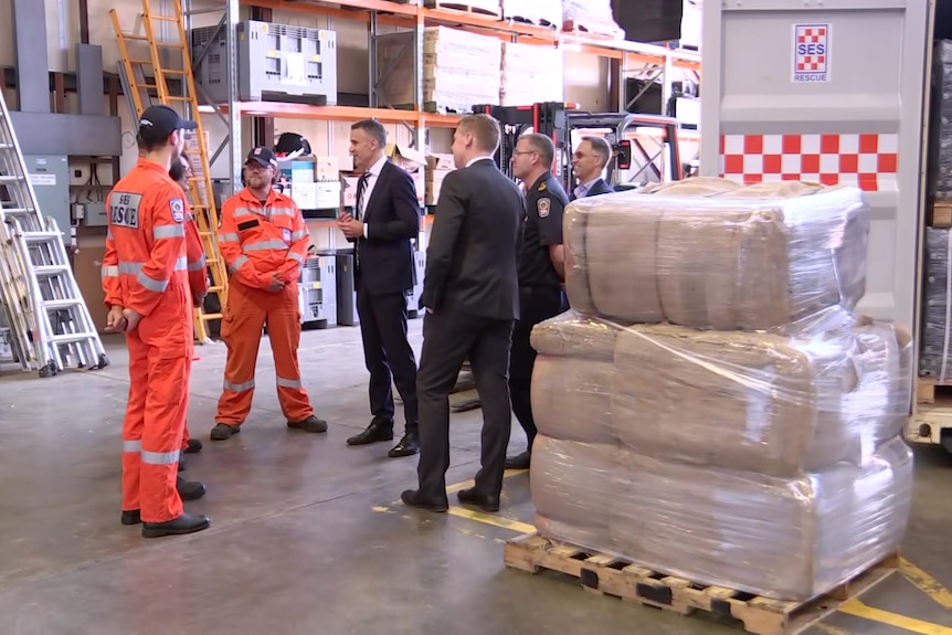 Three men in SES informs talk to Peter Malinauskas and Joe Szackacs who are standing next to a pallet of sandbags