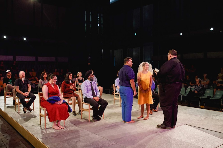 Outdoor stage with people seated on chairs as if at wedding, and bride and groom at front, priest in front. Audience surrounds.