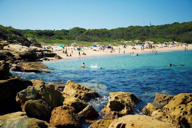 Beachgoers gather on the sand and in the water, with rocks cropping up out of the water in the foreground.