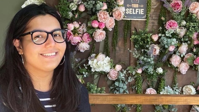 Mina, who has long dark hair and wears glasses, smiles in front of a wall of flowers.