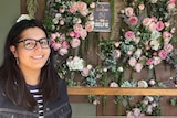 Mina, who has long dark hair and wears glasses, smiles in front of a wall of flowers.