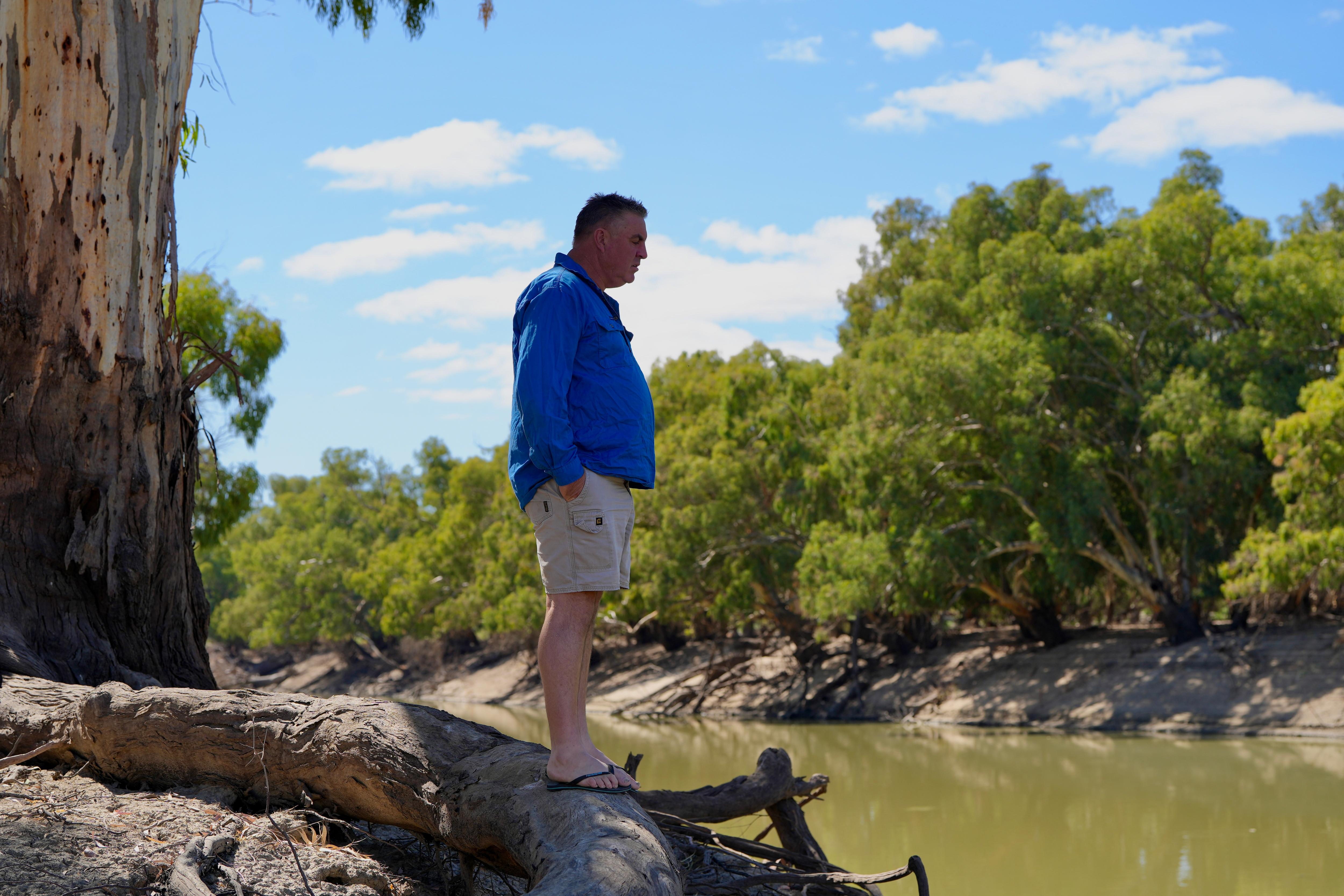 A white man with a blue shirt and beige shorts standing on a tree root looking at the Darling River. 