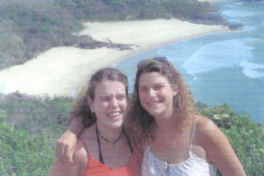Two women hugging with a beach behind them. One in a white shirt one in an orange shirt.