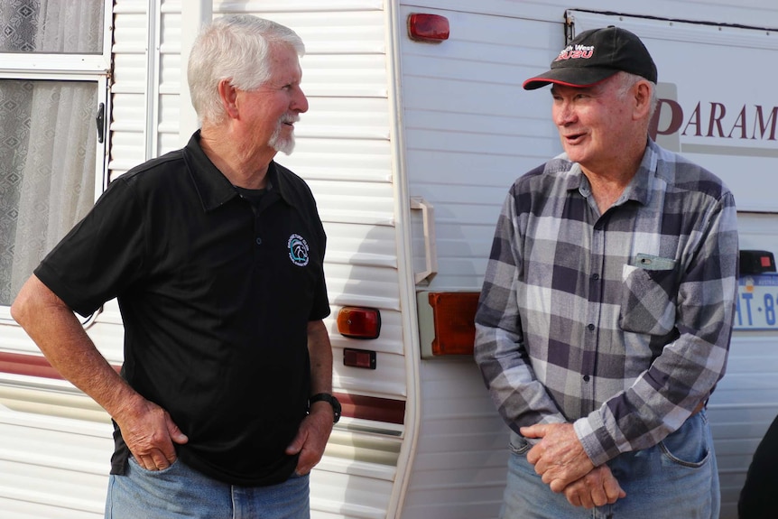 Two older men stand talking to one another alongside a caravan.