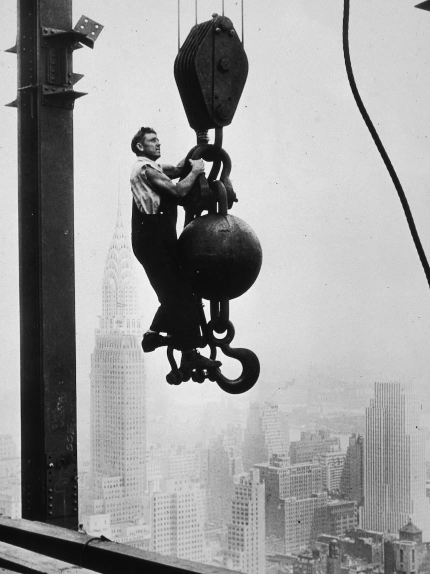A black and white photo of a man hanging from a crane pulley, high above a city.