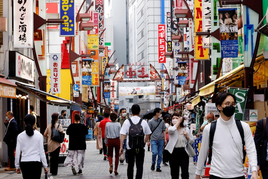 Shoppers move through a busy commericial district with lots of signs hanging from buildings.