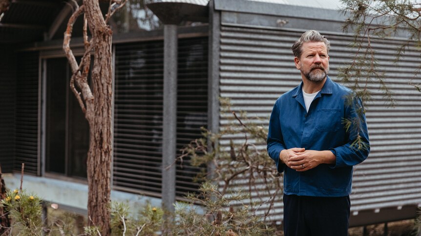 Tim Ross wears a blue long sleeved shirt and stands in front of a house with corrugated iron walls and roof in the bush