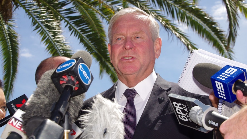 Upshot of Colin Barnett surrounded by microphones under a palm tree.