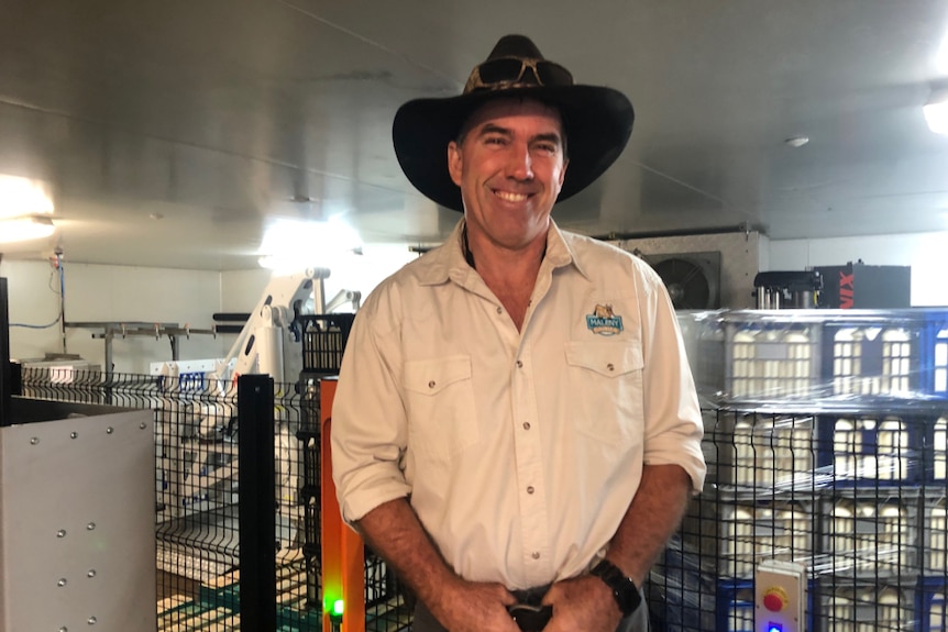 Ross Hopper smiling at the camera with stacked crates of milk and the robot behind him.