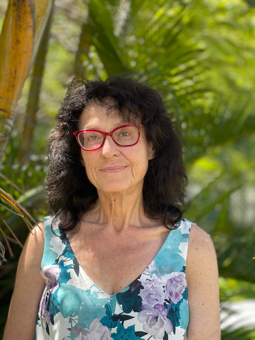 A woman with dark curly hair, wearing red framed glasses and standing against greenery