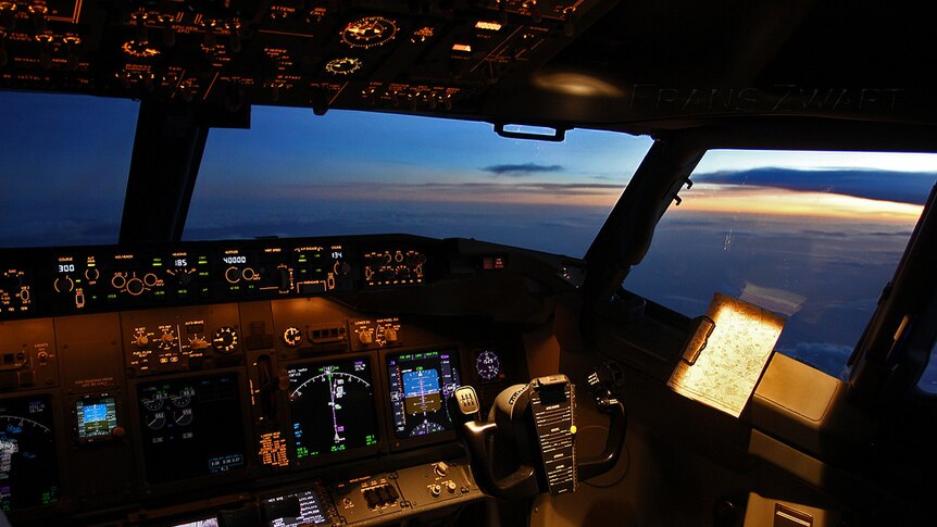 An aircraft cockpit with a number of dimly-lit controls look out to a sky above clouds at sunset.