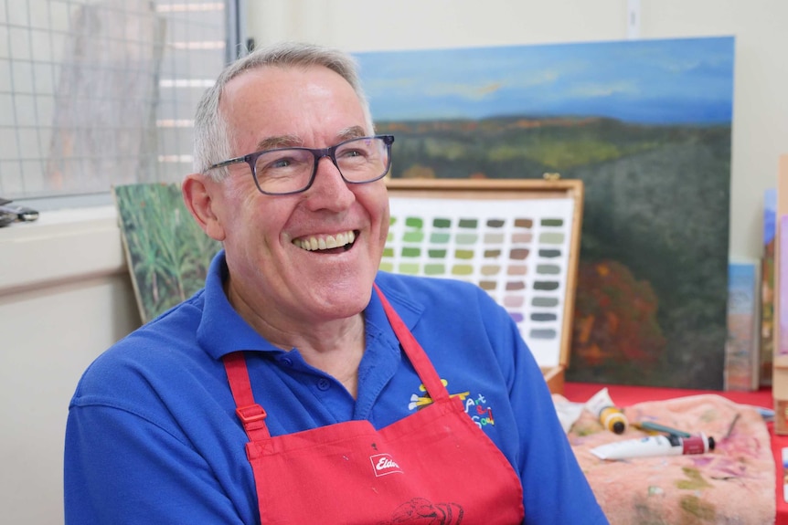 A man with grey hair sits smiling, in front of an art work.