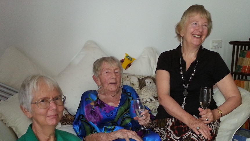 Three women with short hair on a white bed holding glasses