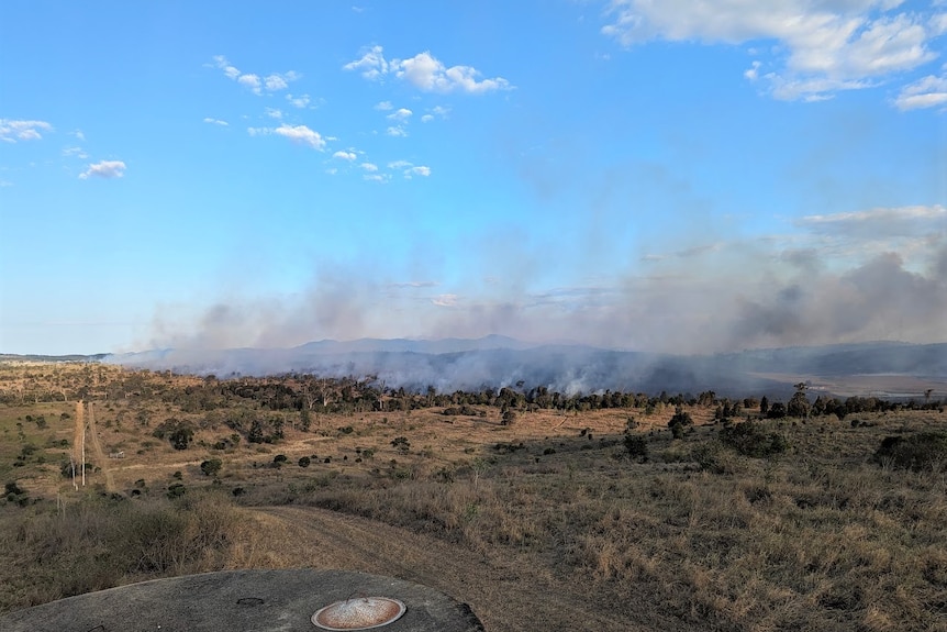 Smoke rising from bushland in front of mountains and a blue sky.