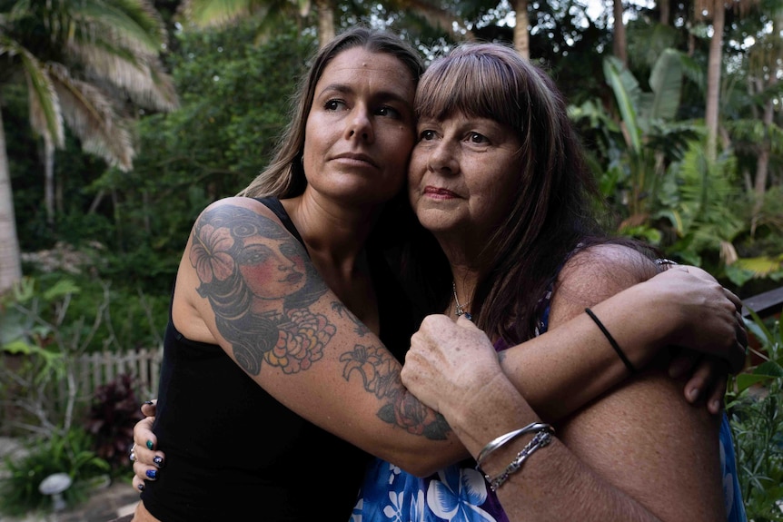A middle aged mother and a daughter in her 30s with tattoos embrace with their arms around each other.