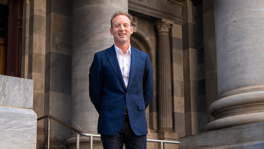 A man wearing a blue suit and white shirt but no tie stands among large classical columns