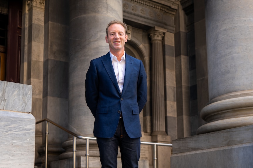 A man wearing a blue suit and white shirt but no tie stands among large classical columns