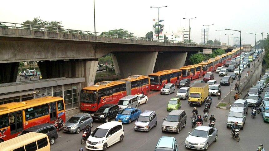 From a high angle, you see a double-decked motorway clogged in car traffic while orange buses are lined up behind each other.