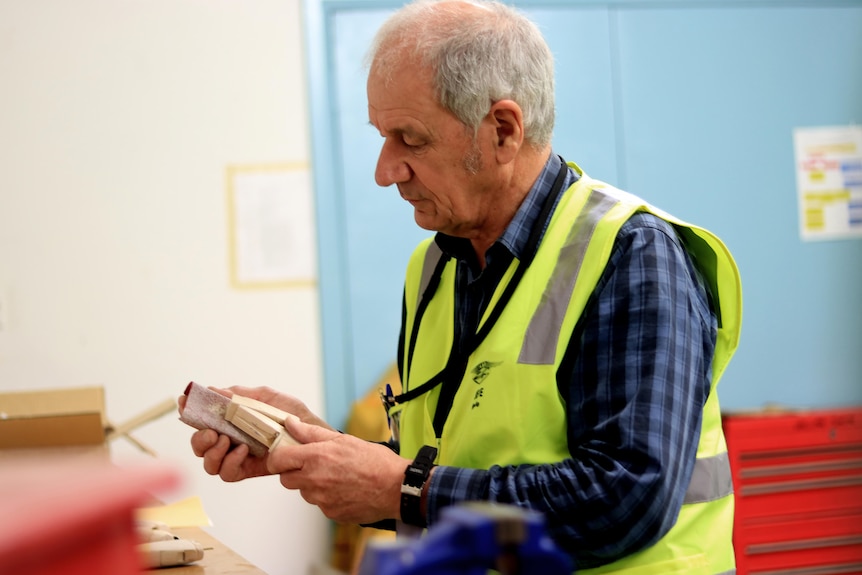 Steve Keddie carves a small piece of wood while wearing a high vis vest and blue shirt in a workshop.