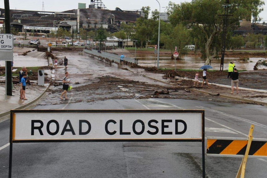 A sign saying "road closed" on a bridge that is flooded by the river beneath it. People with umbrellas are nearby