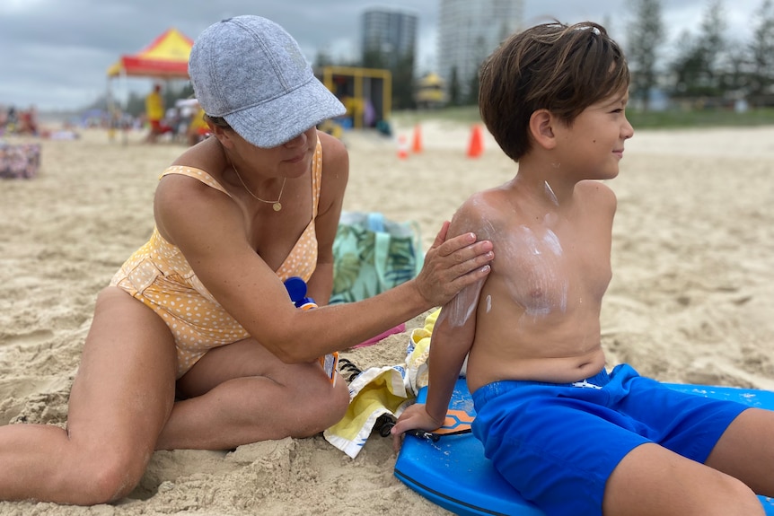 a woman wearing a cap sitting on the sand putting sunblock on a young boy