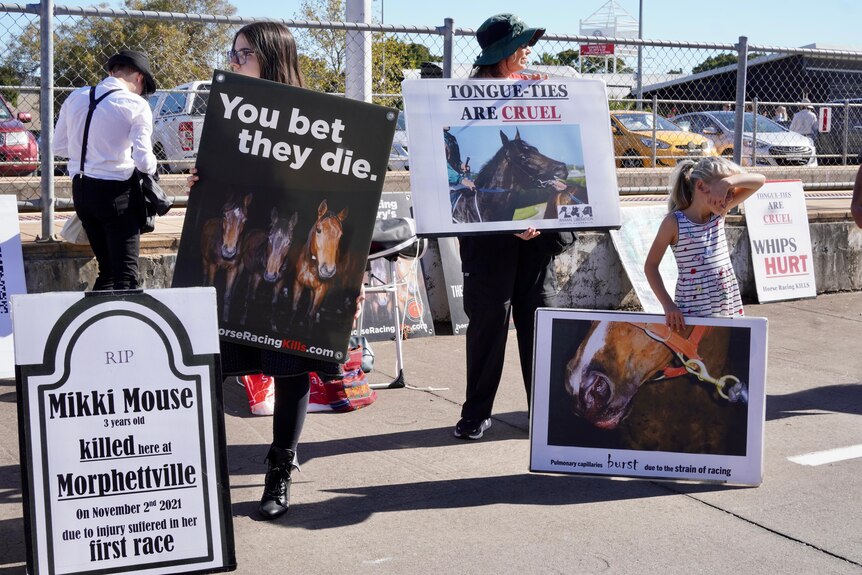 Three protesters stand outside holding signs against horse racing