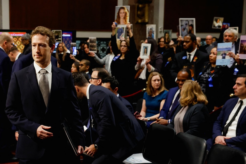 Mark zuckerberg wearing a suit standing in front of a crowd of people holding up pictures on placards