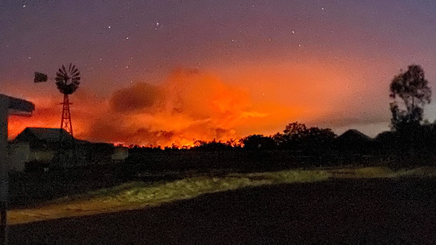 Bushfire burns orange and red on the horizon at night against the silhouette of a windmill, as stars twinkle above in a dark sky