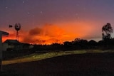 Bushfire burns orange and red on the horizon at night against the silhouette of a windmill, as stars twinkle above in a dark sky