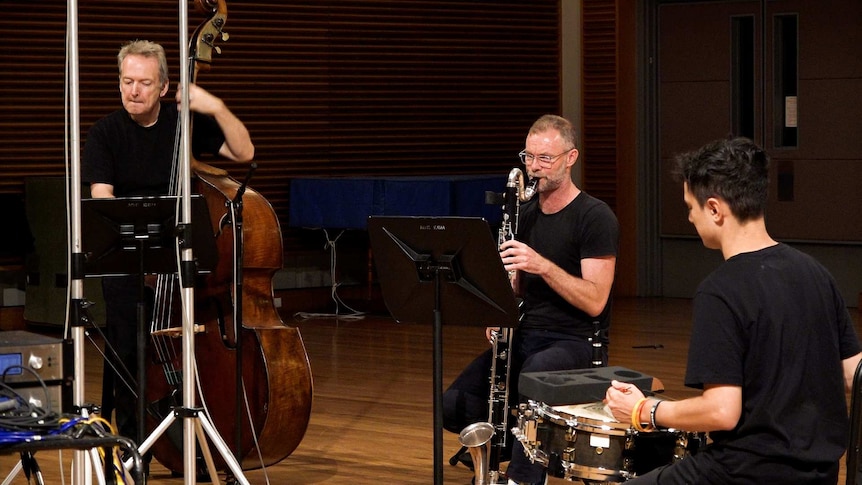 Three musicians in a recording studio playing double bass, bass clarinet, and snare drum.