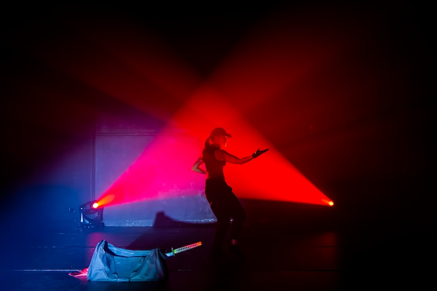 An Asian Australian woman stands on stage, in a cap and gloves. She is illuminated in red light, and has one arm extended.