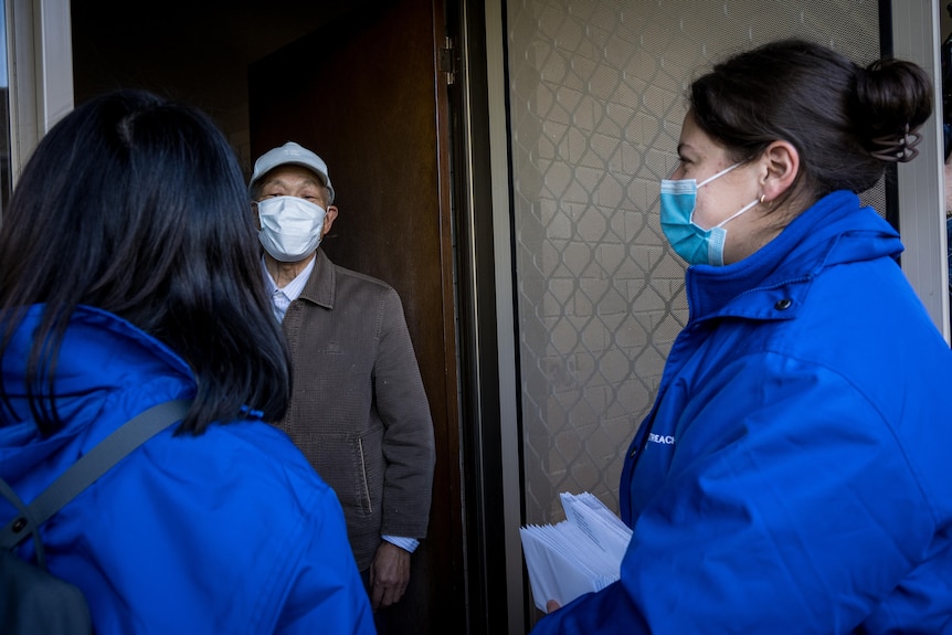 An older man with a mask opens the door to two workers wearing masks and gloves.
