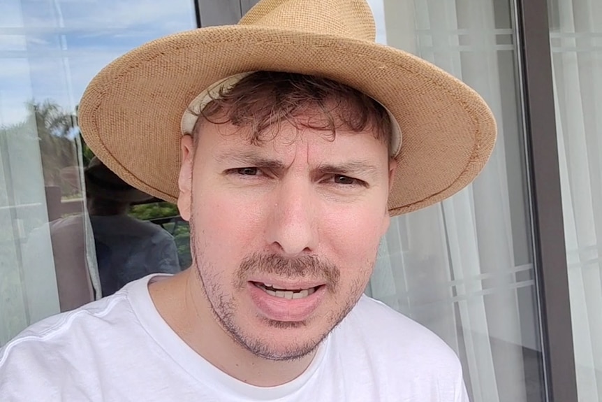 A white man with curly hair wearing brimmed hat and white T-shirt