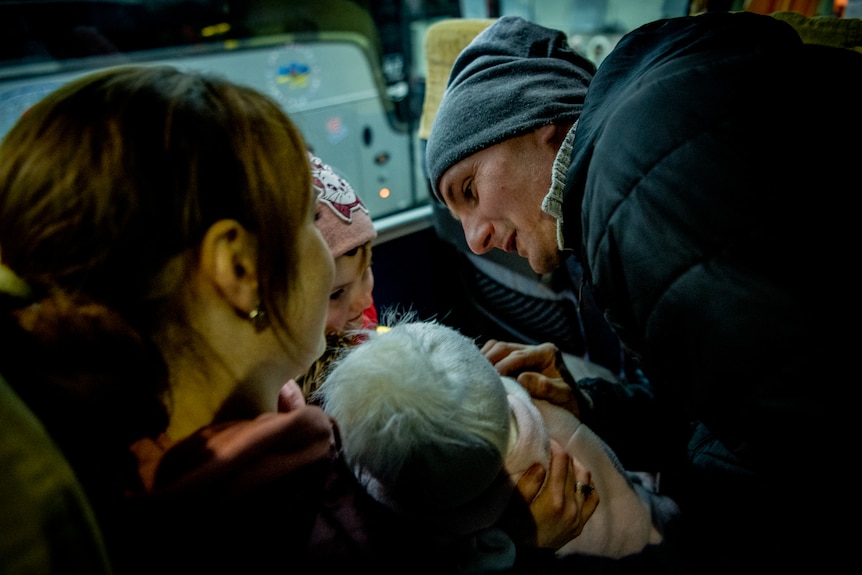 A man leans into a bus seat to kiss his wife and two young children. They are all rugged up 
