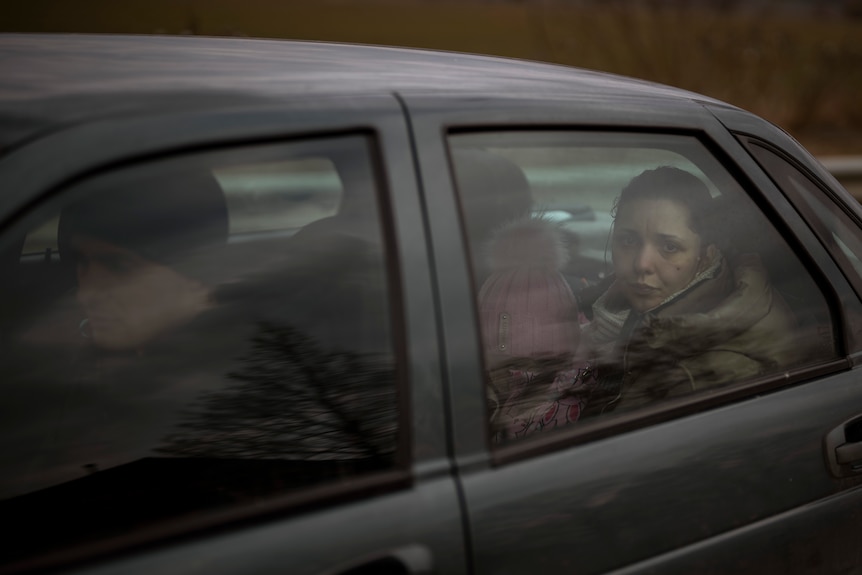 A view of a car as a woman in the back seat looks despondent as she looks out of the window. A child sits next to her.