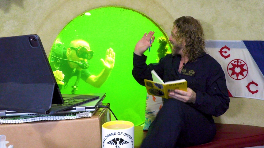 A man in scuba gear waving through a window at a man holding a book.