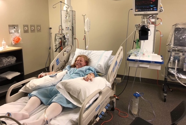 Rebecca Austin lays smiling in a hospital bed, wearing a baby blue gown with two pillows by her side and a tube in her nose.